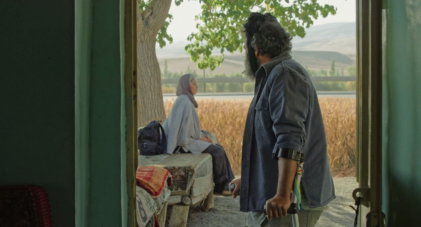 حسن معجونی با عصایی زیر دست ایستاده میان یک در به پانته‌آ پناهی‌ها نگاه می‌کند در نمایی از فیلم جاده خاکی به کارگردانی پناه پناهی