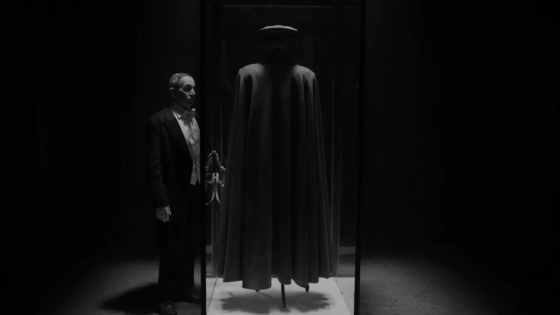 آلفردو کاسترو با لباس پیش‌خدمت کنار یک محفظه‌ی حاوی یک‌دست لباس و شنل نظامی ایستاده است در نمایی سیاه و سفید از فیلم کنت به کارگردانی پابلو لارائین
